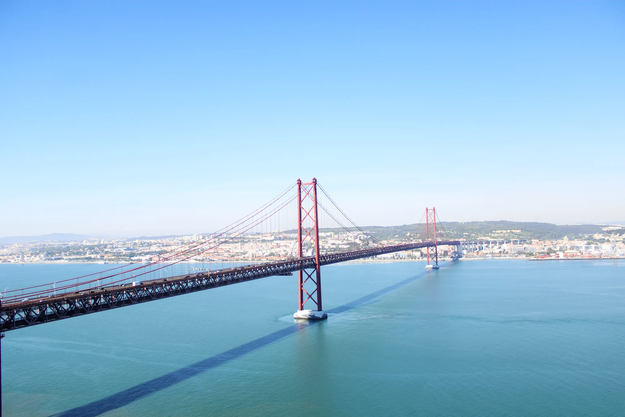 The full Ponte 25 de Abril red suspension bridge with both towers spanning the turquoise Tagus river, with the Lisbon skyline behind under a clear blue sky
