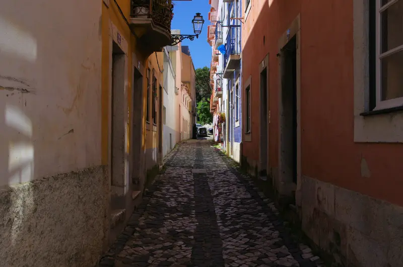 Narrow cobbled calçada lane in Lisbon's Alfama old quarter running between weathered ochre and pink houses with a wrought-iron street lamp and balconies, blue sky beyond