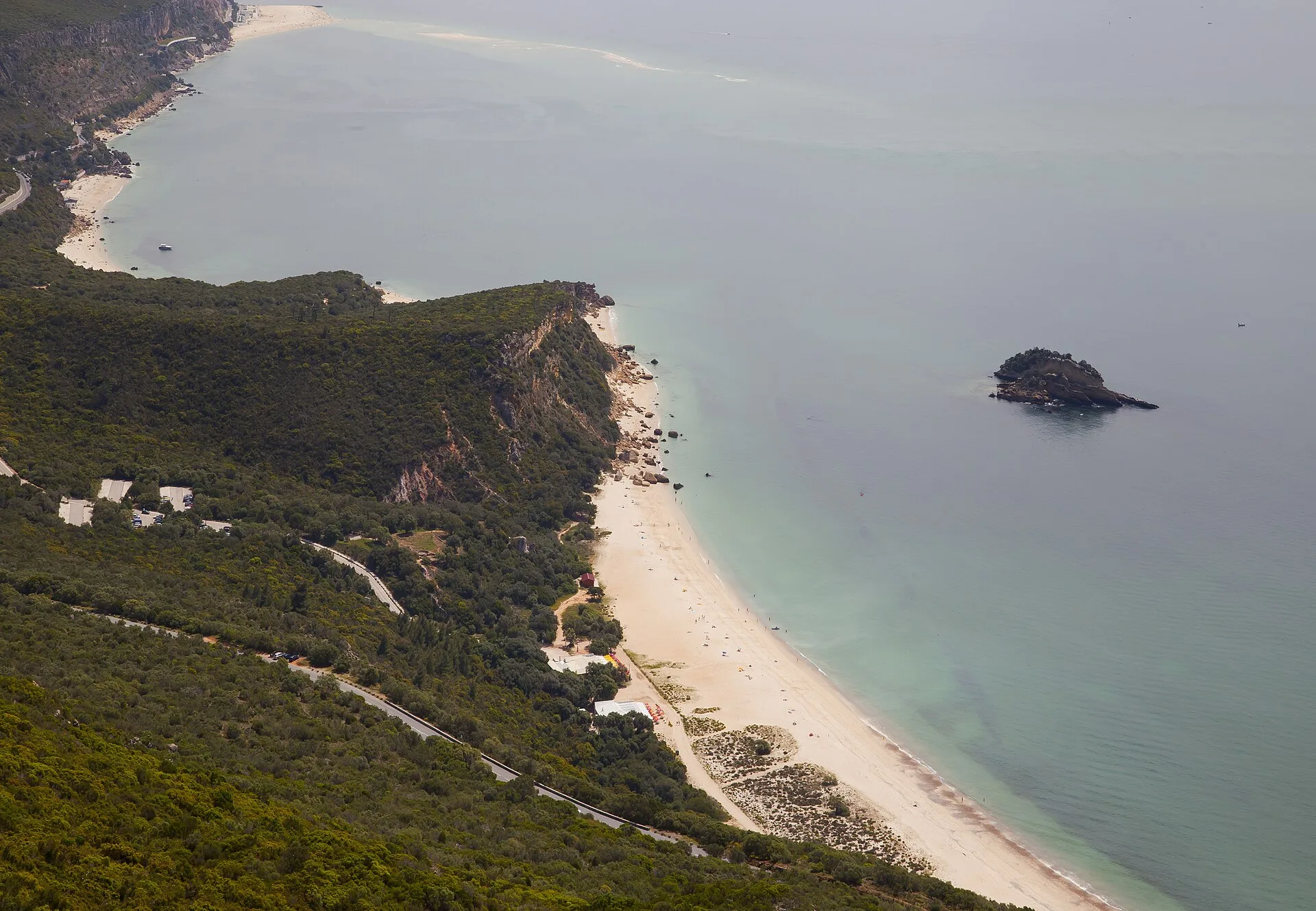 Aerial view of the green wooded hills of Serra da Arrabida meeting a curving white-sand beach and turquoise sea with a small offshore islet, Arrabida Natural Park near Setubal, Portugal