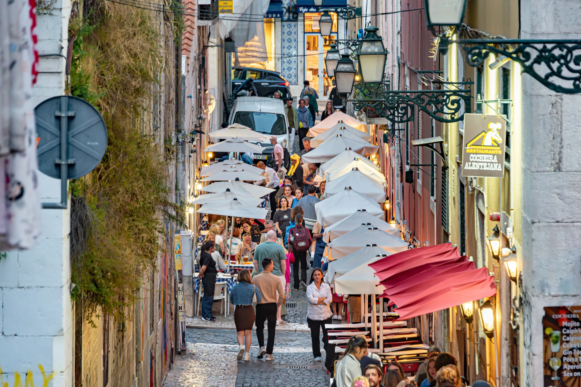 A narrow cobbled street in Bairro Alto, Lisbon, lined with restaurant terraces and umbrellas and crowded with people at dusk, the characteristic nightlife scene of the neighbourhood