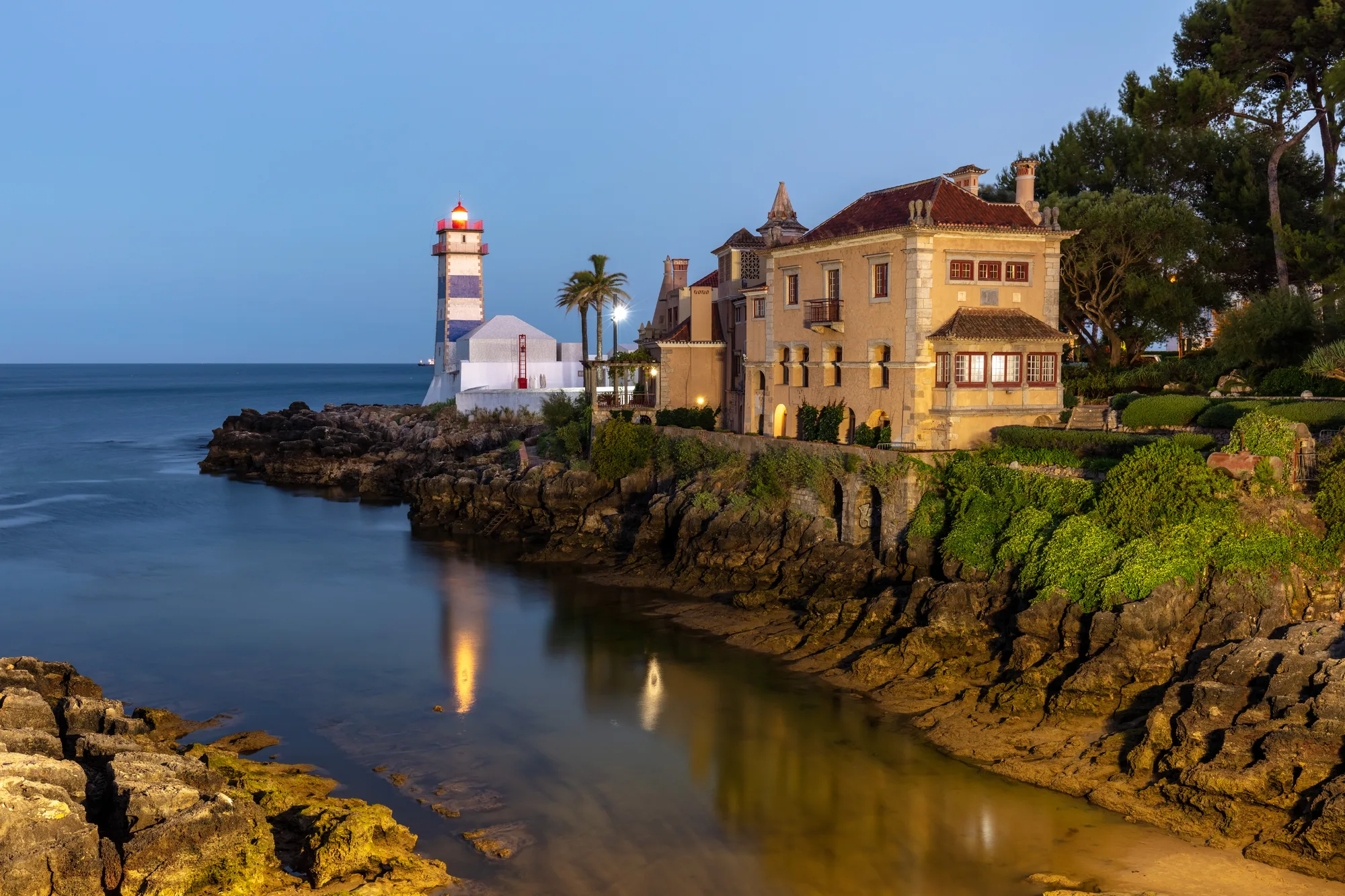 Santa Marta Lighthouse with its blue-and-white striped tower and the seaside museum on the rocky Cascais coastline at dusk, Portugal