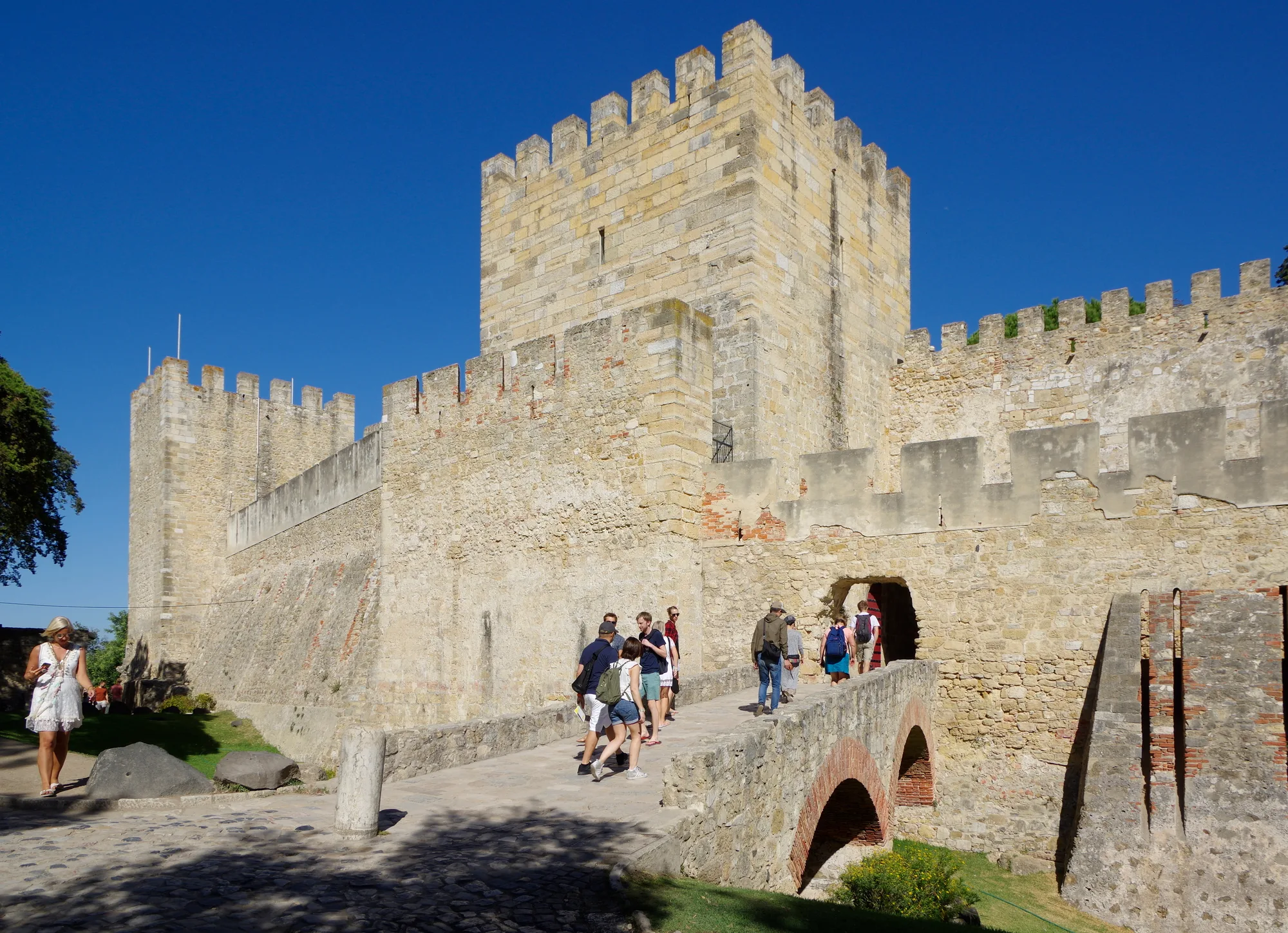Stone crenellated ramparts and towers of Castelo de São Jorge, the hilltop castle in Lisbon, with the entrance bridge and visitors in the foreground under a blue sky