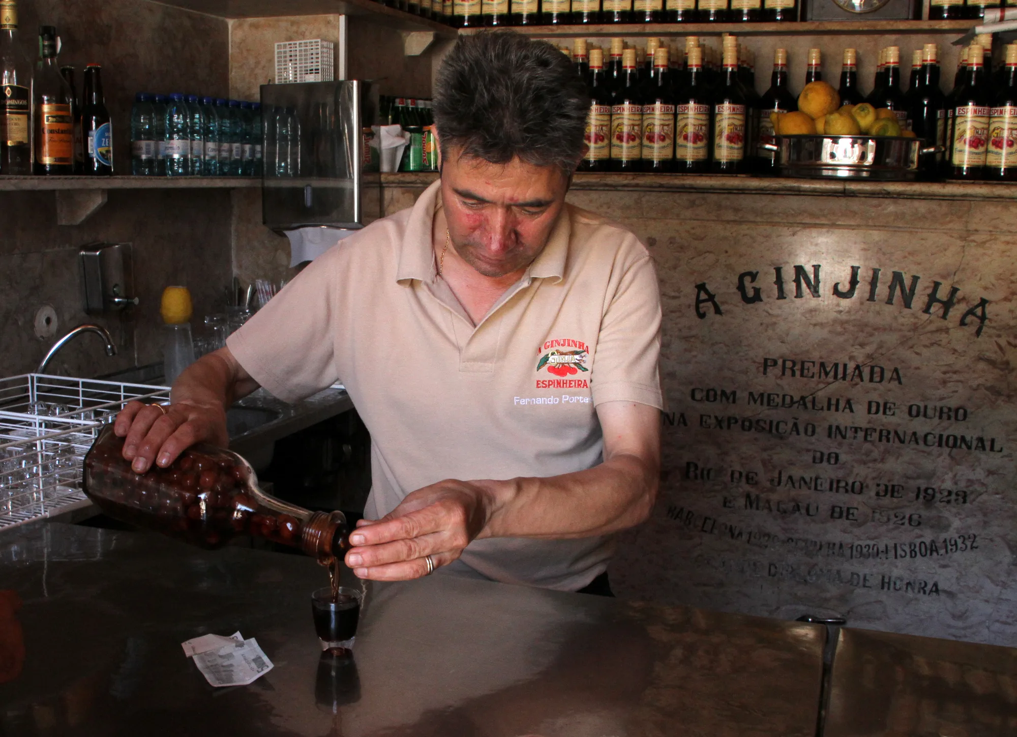 Barman pouring dark red ginjinha sour-cherry liqueur from a bottle into a small glass at the famous A Ginjinha (Espinheira) bar in Lisbon, marble wall engraved A GINJINHA behind