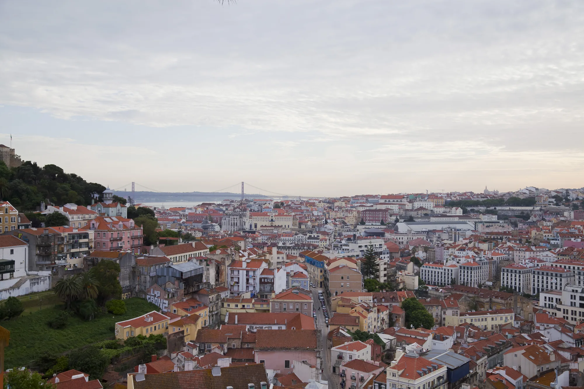 City panorama from Miradouro da Graça in Lisbon: a sea of red-tiled rooftops with the green Castelo de Sao Jorge hill on the left and the 25 de Abril bridge over the Tagus in the distance