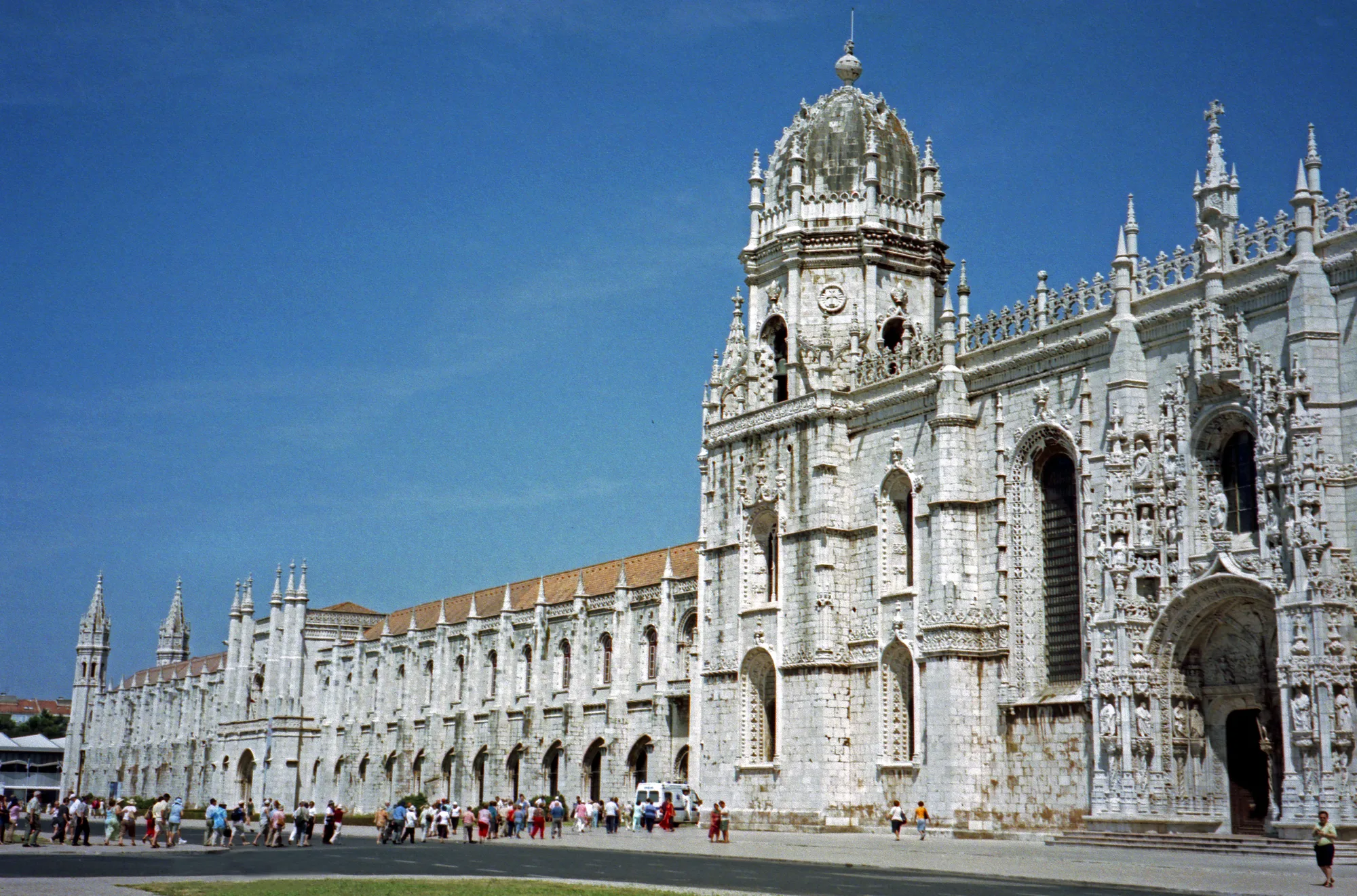 The long ornate Manueline white-limestone south facade of Jerónimos Monastery in Belém, Lisbon, with its carved south portal, central tower and the church wing stretching to the left under a clear blue sky