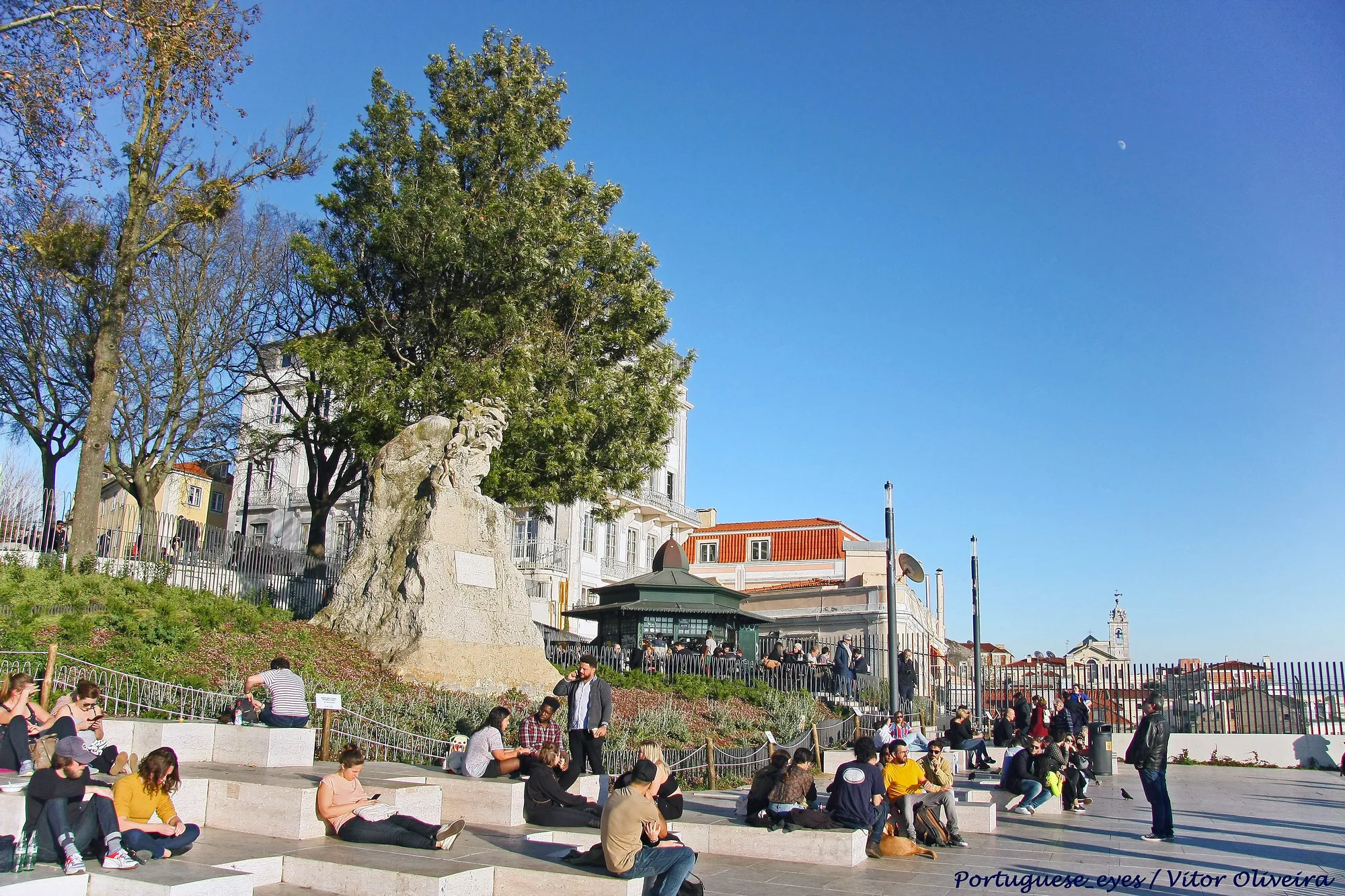 Miradouro de Santa Catarina (Adamastor) terrace in Lisbon on a clear day, with the weathered Adamastor stone monument and crowds of people sitting on the stepped terrace and kiosk