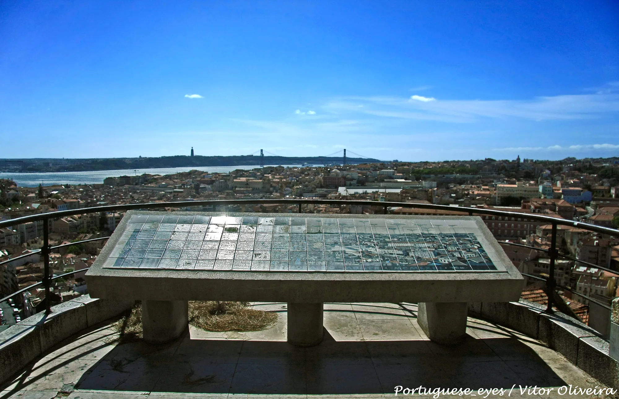 Panorama from Miradouro da Senhora do Monte in Lisbon: a tiled orientation table on the terrace overlooking the city rooftops, the Tagus river and the 25 de Abril bridge under a blue sky