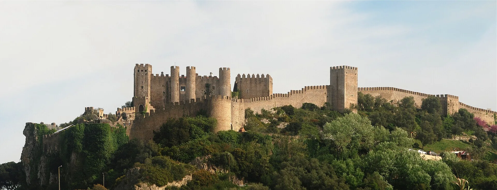 The medieval castle and crenellated stone walls of Obidos crowning a green hilltop in warm light, Portugal