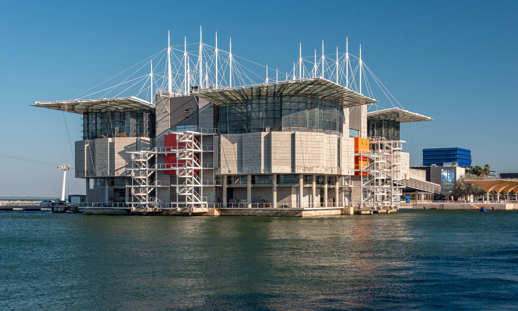 The Oceanário de Lisboa building standing on the water in Parque das Nações, Lisbon, with its mast-and-cable roof reflected in the marina