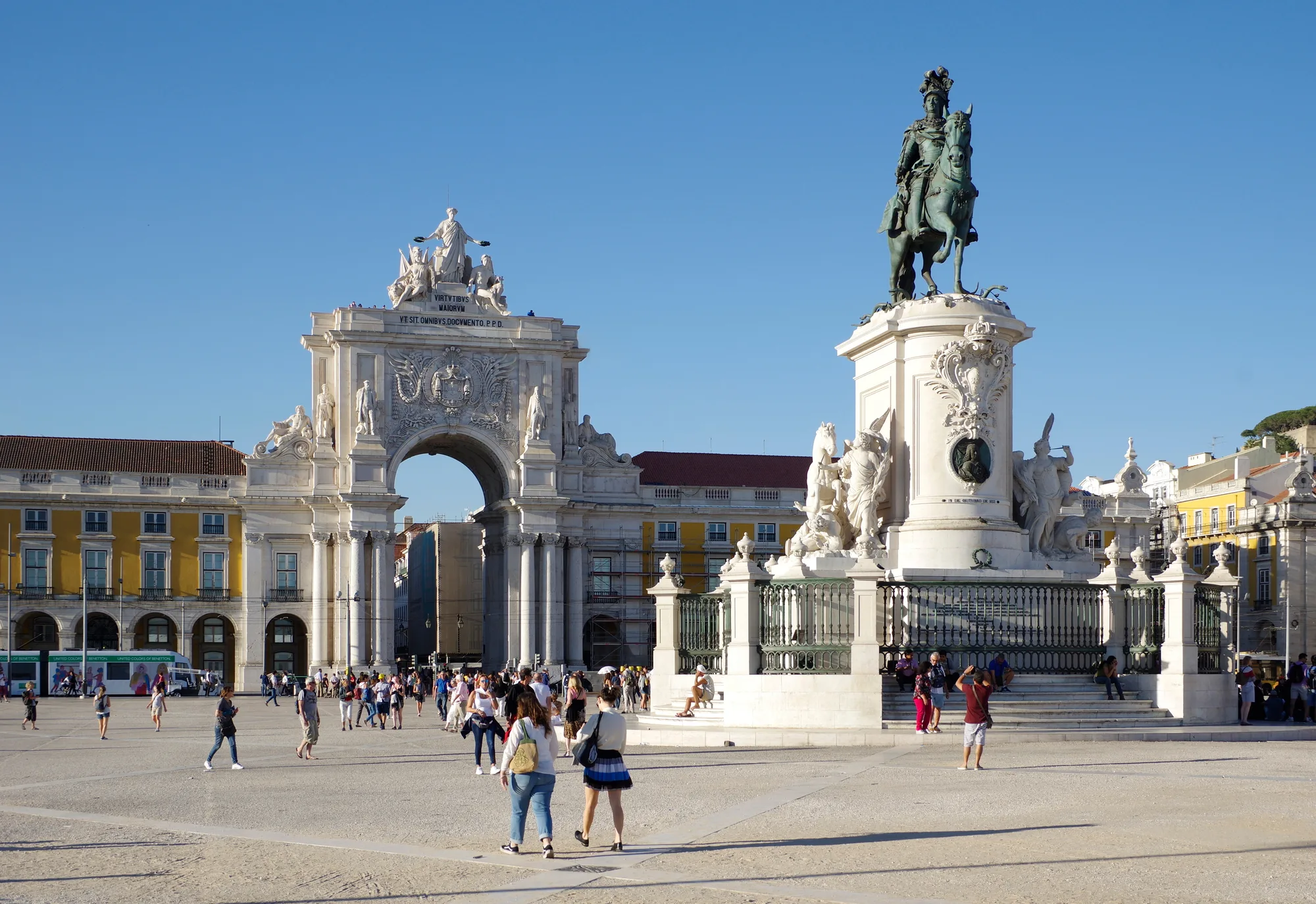Praça do Comércio in Lisbon: the bronze equestrian statue of King José I, the white Arco da Rua Augusta triumphal arch behind it, and the yellow arcaded riverfront buildings, with people crossing the square under a blue sky