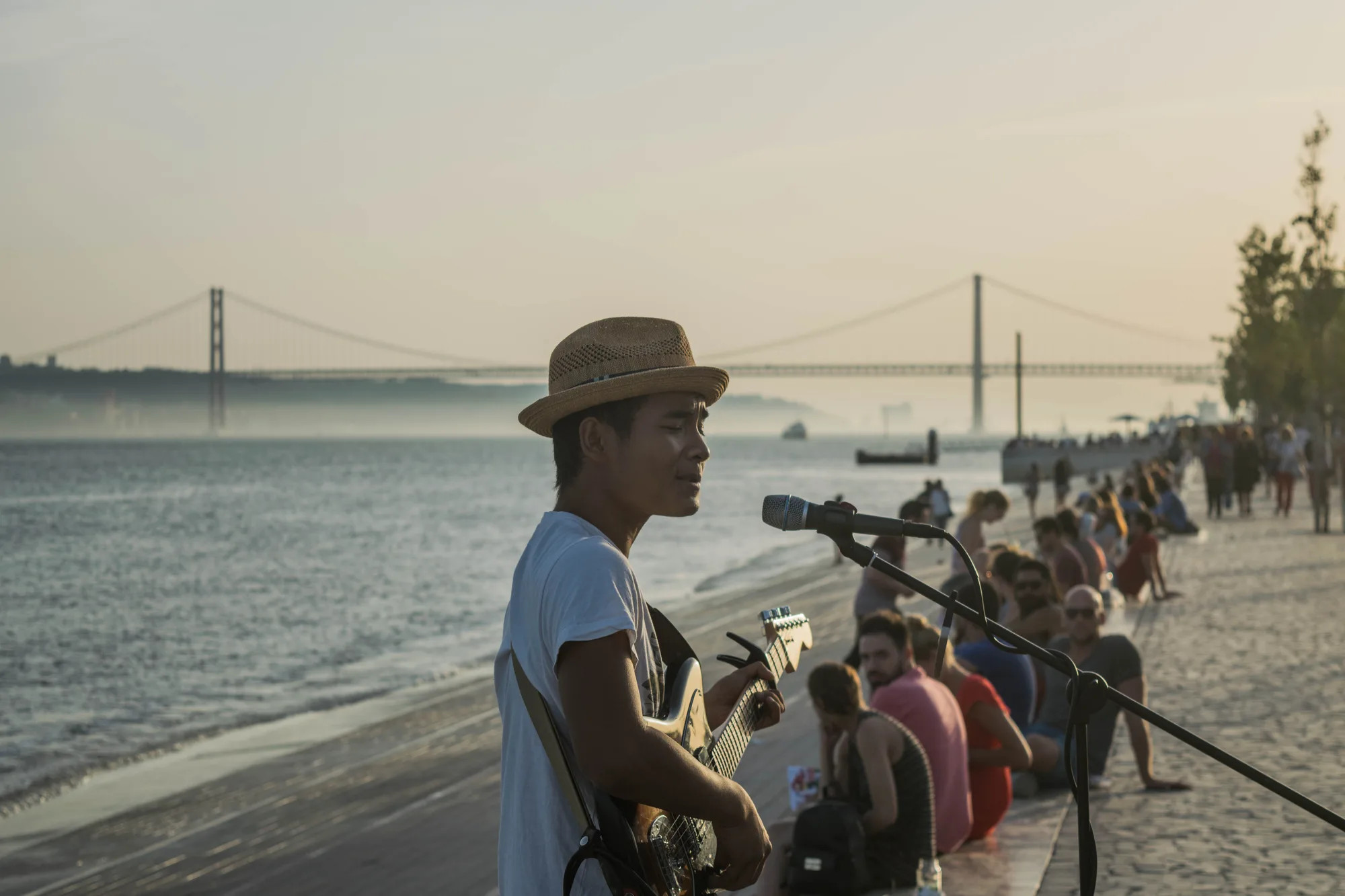 The Ribeira das Naus riverfront promenade in Lisbon at golden hour, with a street musician playing guitar and crowds sitting on the sloping steps into the Tagus, the 25 de Abril bridge in the background
