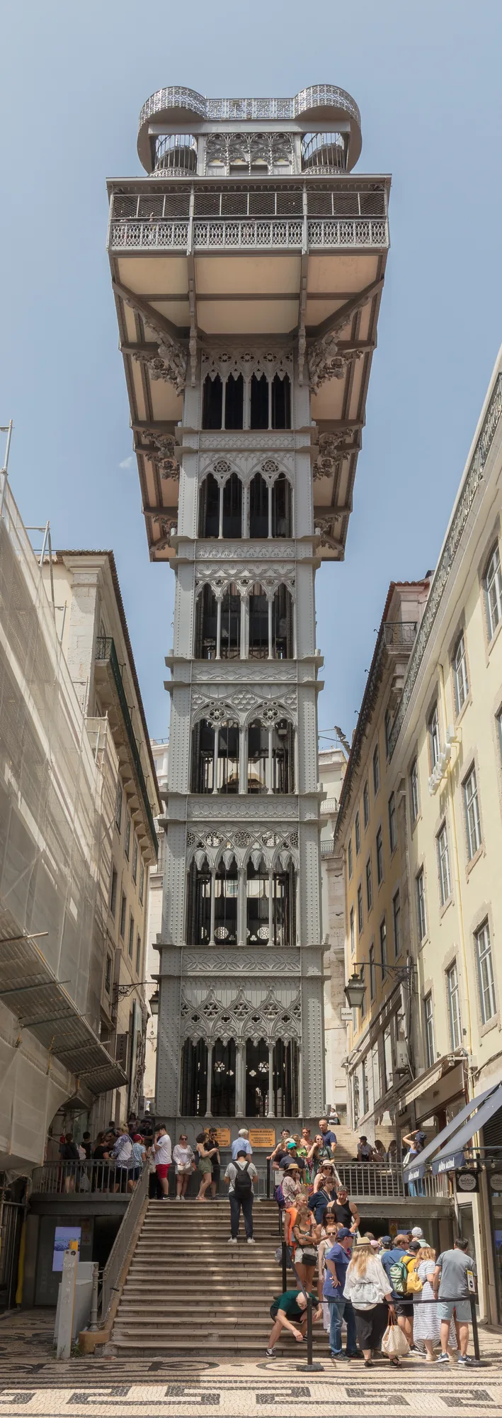 The full wrought-iron neo-Gothic tower of the Santa Justa Lift (Elevador de Santa Justa) rising between buildings in the Baixa district of Lisbon, with visitors on the cobbled steps at its base