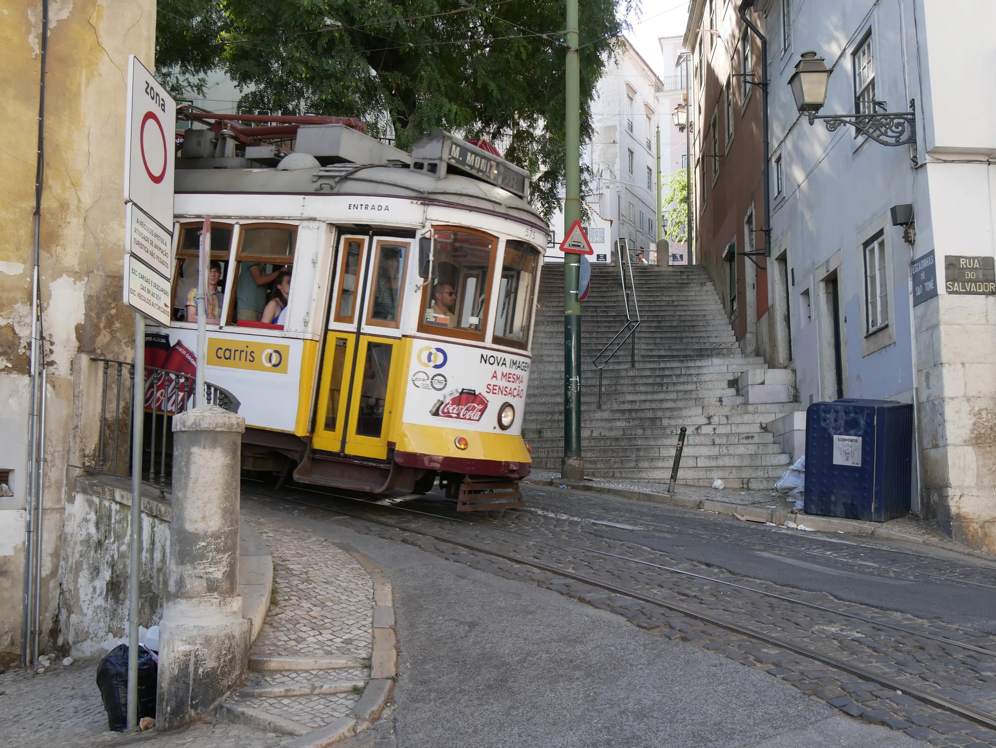 Classic yellow and white Remodelado Tram 28 (Eléctrico 28) on a steep cobbled Lisbon street beside a staircase and old buildings in the historic quarter